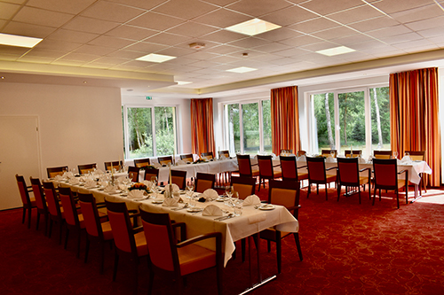 Ein großer, elegant eingerichteter Speisesaal mit langen Tischreihen, die für ein formelles Essen gedeckt sind. Große Fenster mit orangefarbenen Vorhängen lassen Tageslicht herein und bieten Blick auf grüne Bäume.