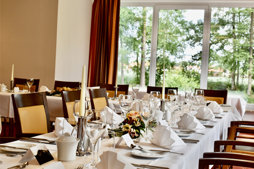 Elegantes Restaurant mit gedeckten Tischen, geschmückt mit weißen Servietten und Gläsern, umgeben von großen Fenstern mit Blick auf grüne Bäume.