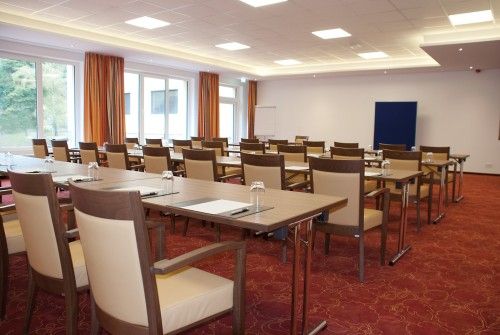 Moderner Konferenzraum mit Tischreihen, Stühlen und Schreibmaterialien auf rotem Teppichboden, umgeben von großen Fenstern.