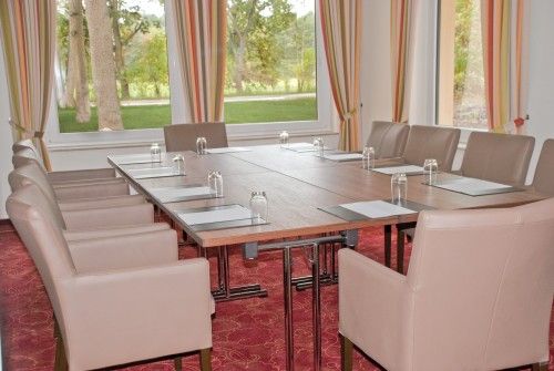 Konferenzraum mit einem rechteckigen Tisch, umgeben von beigen Stühlen, ausgestattet mit Schreibunterlagen und Wassergläsern, bei Tageslicht durch große Fenster.