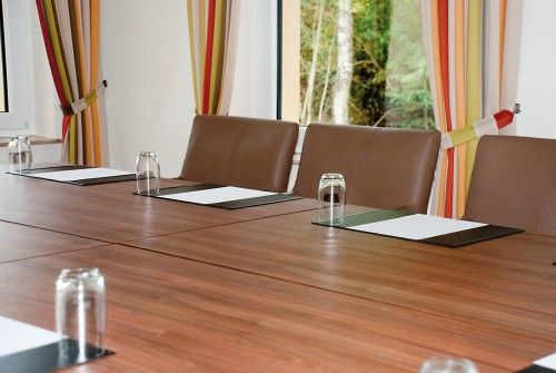 Ein leerer Konferenztisch mit braunen Stühlen, Gläsern und Schreibunterlagen, vor einem Fenster mit gestreiften Vorhängen.