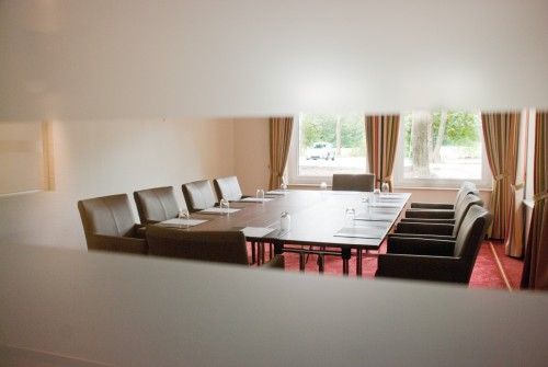 Konferenzraum mit einem langen Tisch, umgeben von schwarzen Stühlen und Fenstern im Hintergrund.