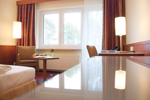 Gut beleuchtetes Hotelzimmer mit modernem Dekor, einem bequemen Sessel, Tisch mit Obst und Blumen, Schreibtisch, und großen Fenstern.