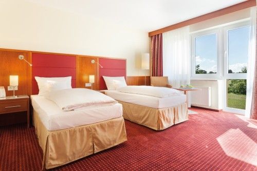 Helles Hotelzimmer mit zwei Einzelbetten, rotem Teppich und großem Fenster mit Blick nach draußen.