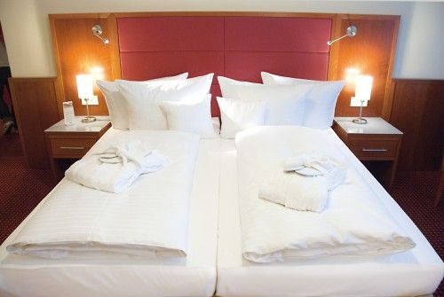 Doppelbett in einem Hotelzimmer, mit roten Kopfteilen, zwei Nachttischen und Leselampen. Auf jedem Bett befindet sich ein gefalteter Bademantel.