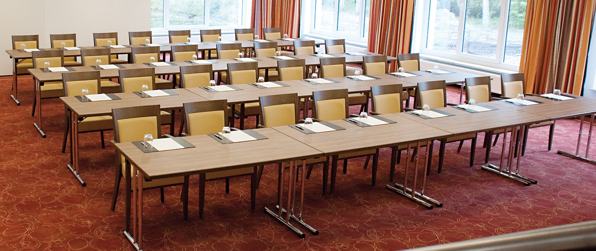Konferenzraum mit rechteckigen Tischen und Stuhlreihen auf Teppichboden, ausgestattet für ein Meeting.