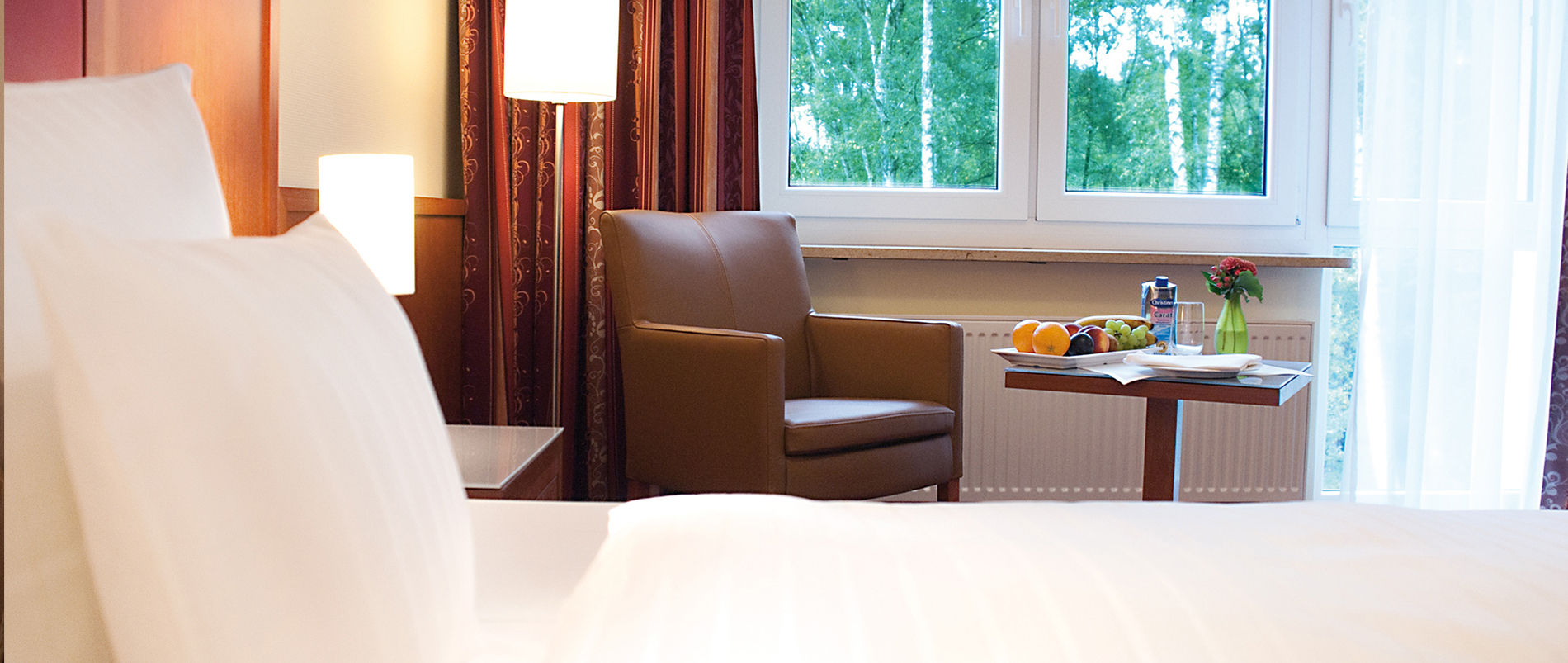 Gemütliches Hotelzimmer mit Sessel, Tisch und einem großen Fenster mit Blick ins Grüne.